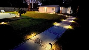 Um quintal com um caminho pavimentado, um pequeno galpão e uma casa.