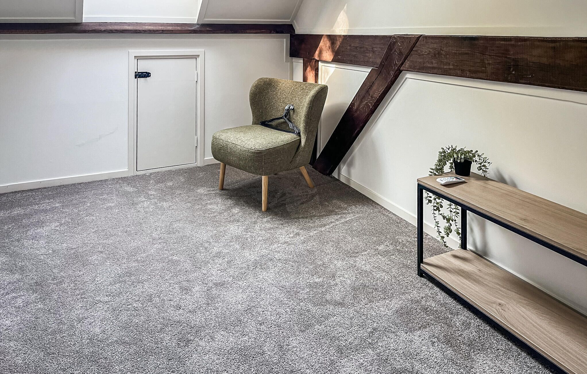 Una habitación con alfombra gris, un banco de madera, una mesita con una planta y una puerta.