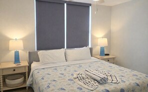 Una habitación de hotel con una cama, dos mesitas de noche, un ventanal con cortinas y una lámpara colgante.