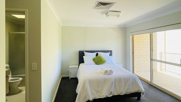 A bedroom with a large bed, a small bedside table, a window with a view, and a door leading to a bathroom. - Rehoboth homes (The Vines)