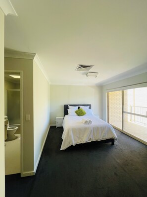 A bedroom with a large bed, a small bedside table, a window with a view, and a door leading to a bathroom. - Rehoboth homes (The Vines)