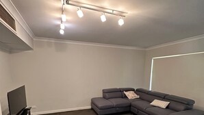 A modern living room with a sectional sofa, a flat-screen TV, and a ceiling light fixture. - Rehoboth homes (The Vines)