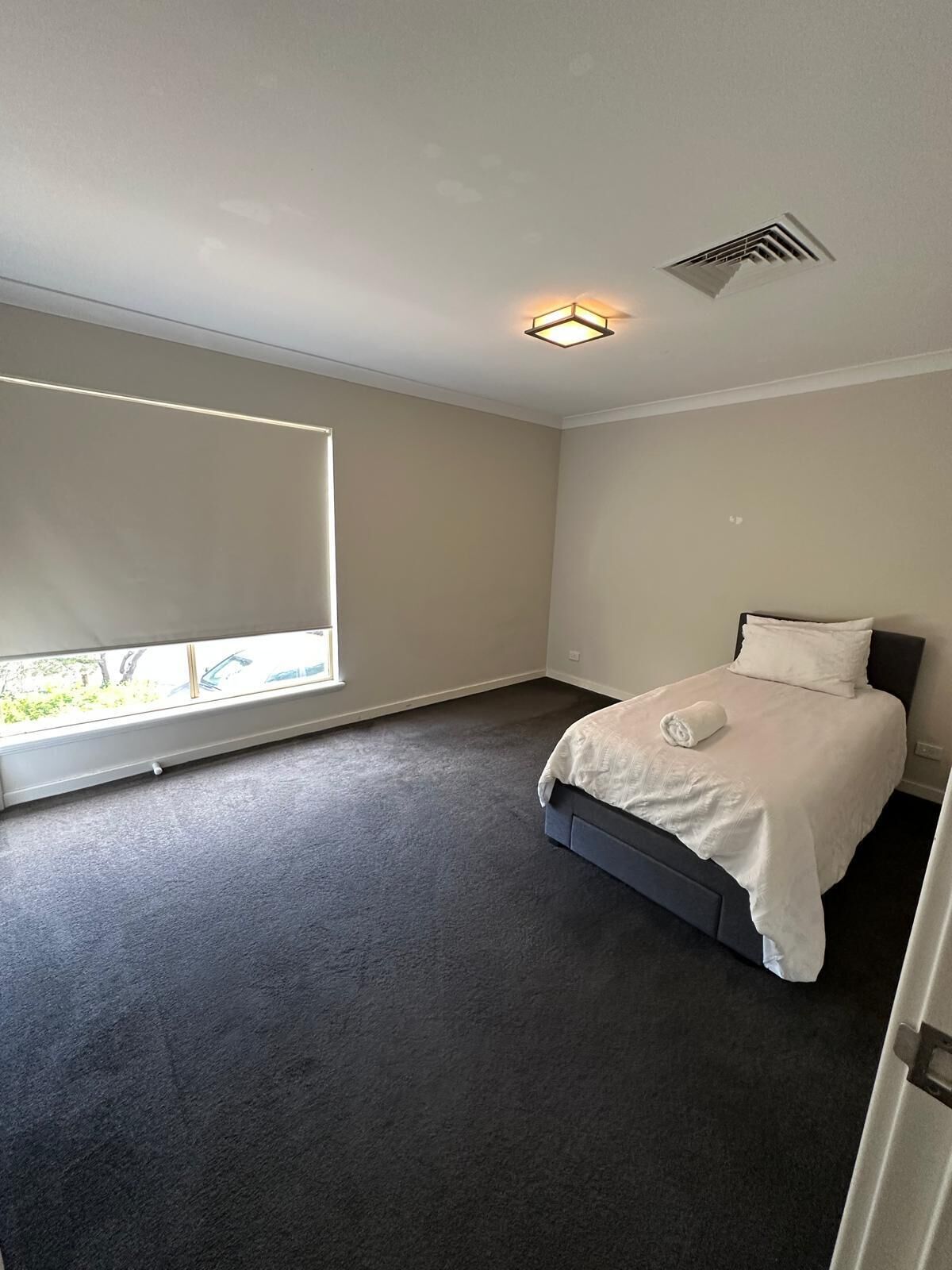 A bedroom with a large window, a bed with a white comforter, and a ceiling vent.