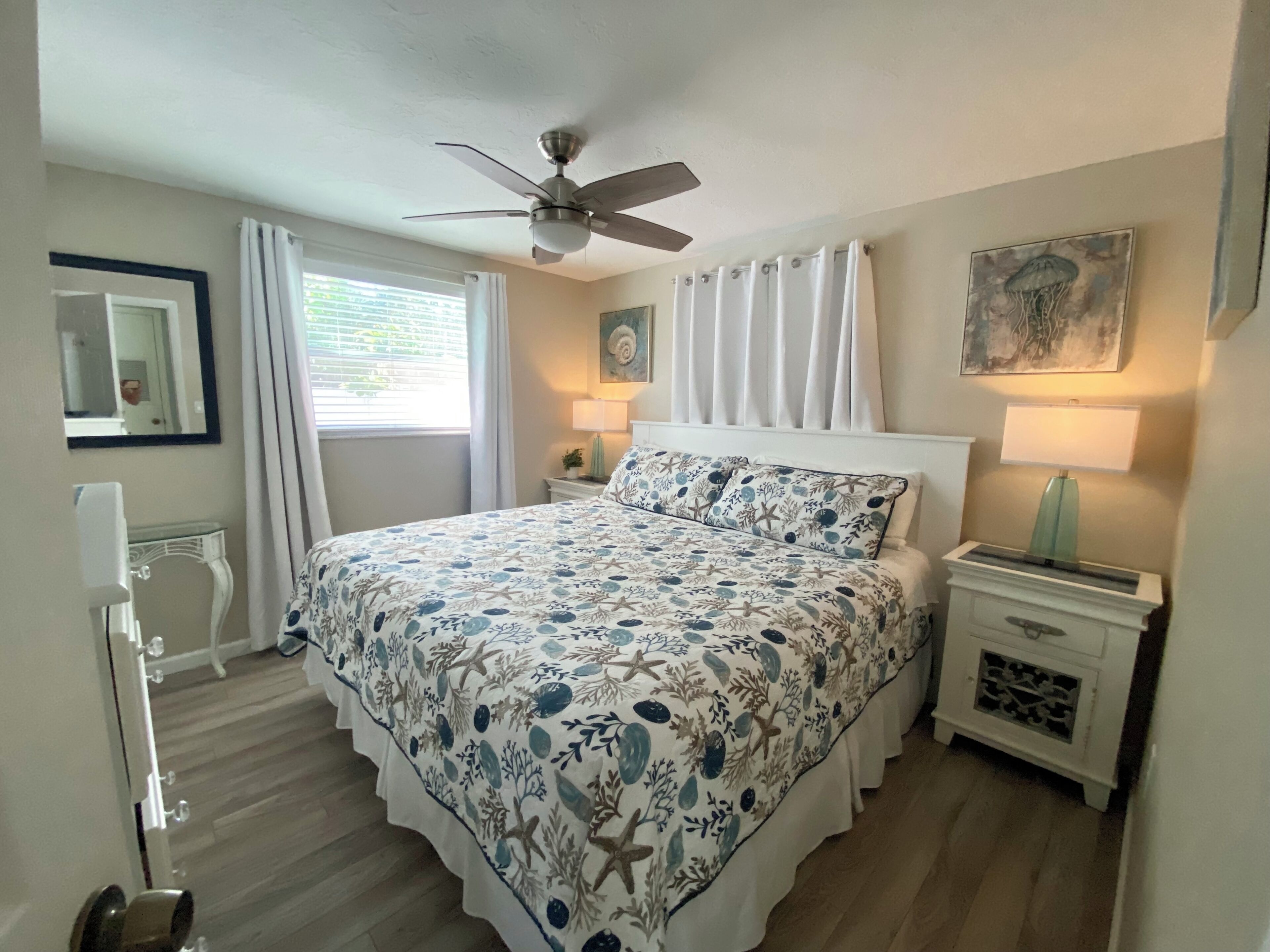 A bedroom with a bed, two bedside tables, a ceiling fan, a window with curtains, and a wall-mounted lamp.