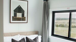 A bedroom with a bed, a framed picture on the wall, a window with a view of the countryside, and an exit sign.