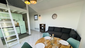 A dining area with a round wooden table set for four, surrounded by teal chairs. A black sofa is against the wall, and a ladder is visible in the background leading to a kitchen area with light green cabinets.