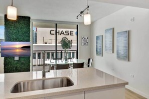 A modern kitchen with a stainless steel sink, a dining area with a glass table, and a view of a cityscape through a large window.
