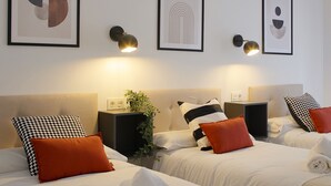 Ein Hotelzimmer mit zwei Betten, jeweils mit weißer Bettwäsche und orangenen Kissen. An der Wand befinden sich gerahmte abstrakte Kunstwerke und Wandlampen.