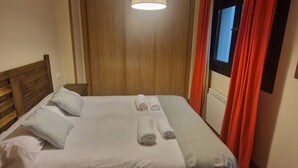 Una cama bien hecha con almohadas y toallas enrolladas, un cabecero de madera, una lámpara colgante y una ventana con cortinas rojas.