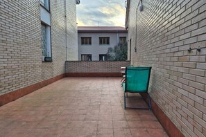 Un patio con piso de baldosas, una silla verde y un edificio con tejado rojo al fondo.