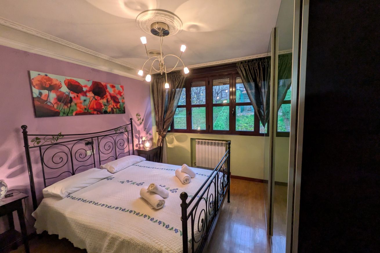 A bedroom with a bed, a nightstand, a painting, a window with curtains, and a radiator.