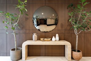 A wooden wall with a round mirror reflecting a room, a wooden console table with vases, and two potted plants. - Kins Hotel (Los Angeles)