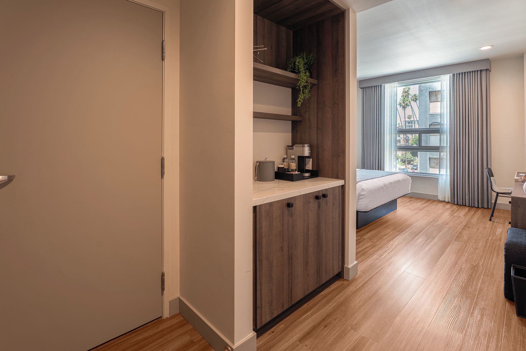 Una habitación de hotel moderna con cama, escritorio, silla, ventana con cortinas y una planta en un estante.