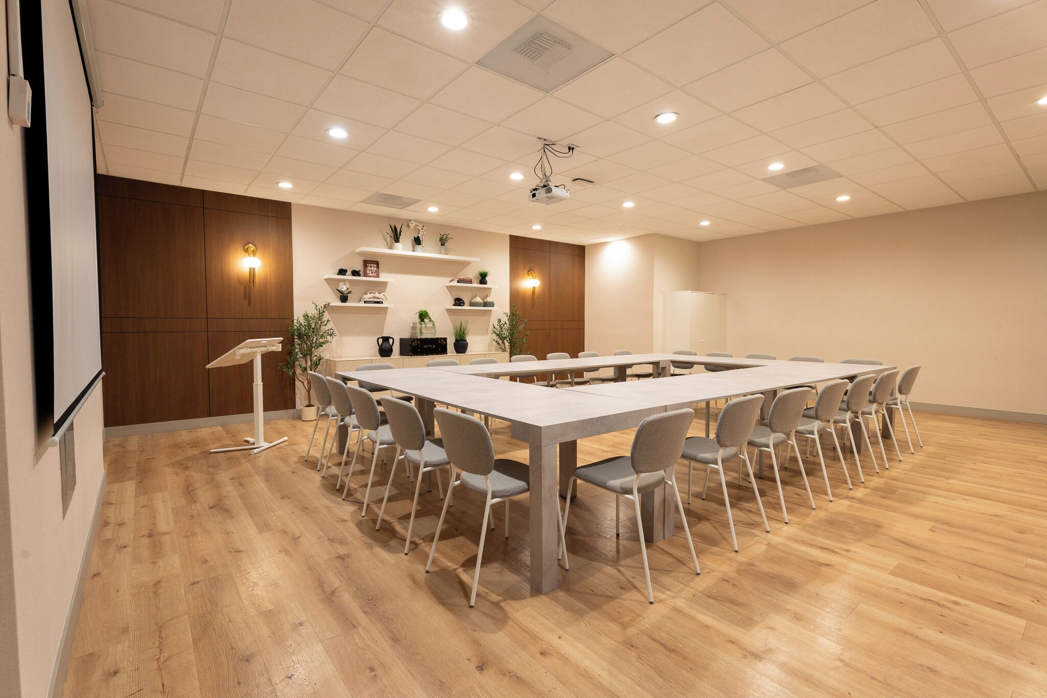 A conference room with a long table, chairs, a podium, and shelves with plants.