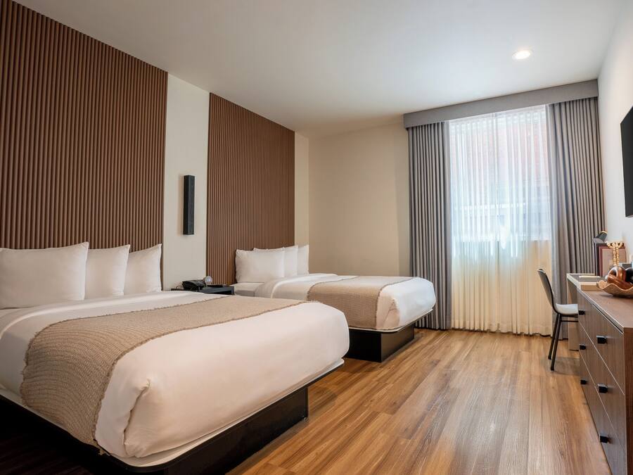 A hotel room with two beds, a desk, a chair, a TV, and a large window with curtains.