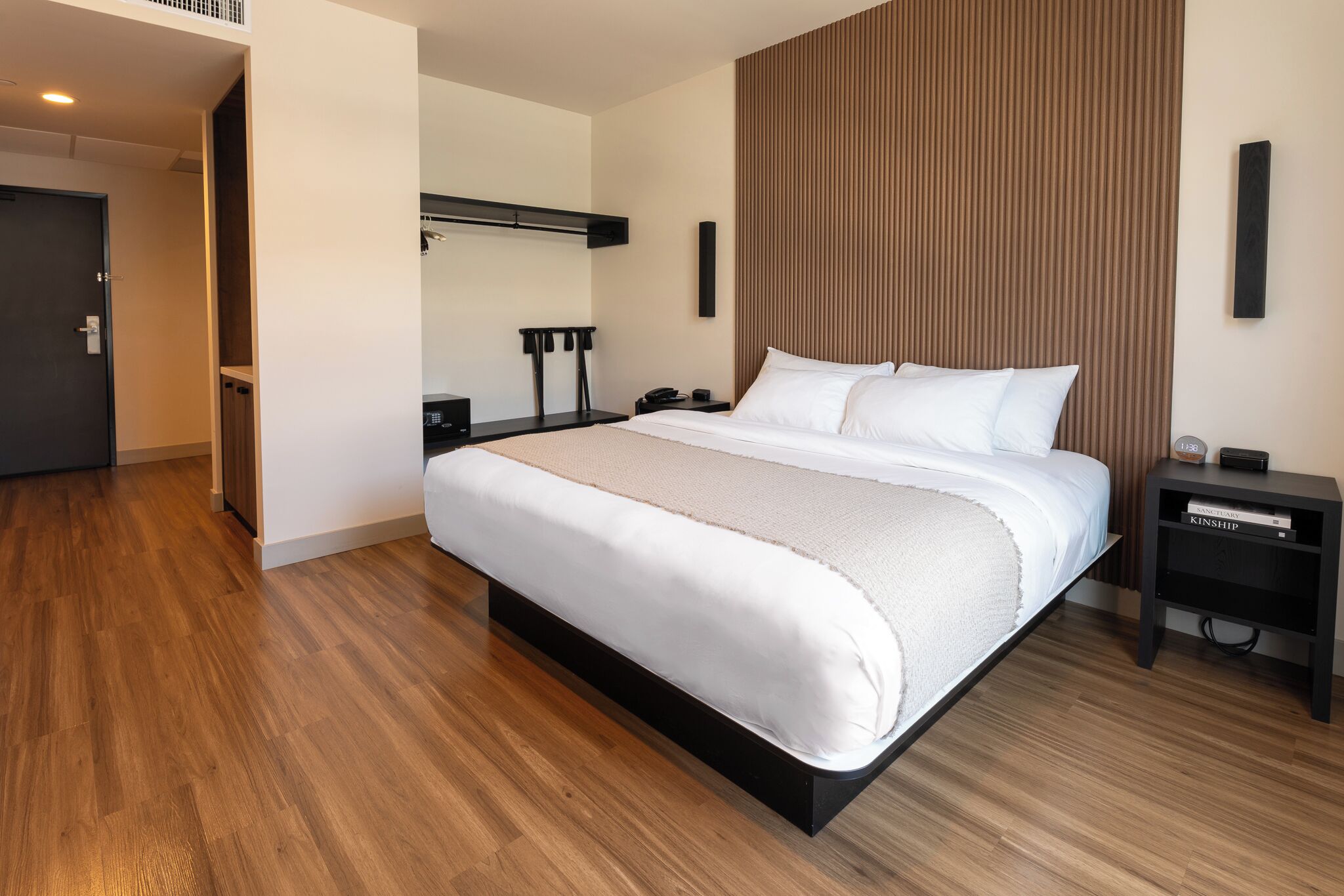 A modern hotel room with a large bed, a bedside table with a lamp, a wall-mounted shelf, and a wooden floor.