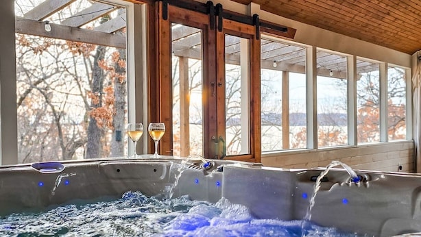 A hot tub with two glasses of wine, wooden beams, and a view of trees through large windows. - Lakefront Oasis | Hot Tub | Game Room | 3+ Acres (Stigler)