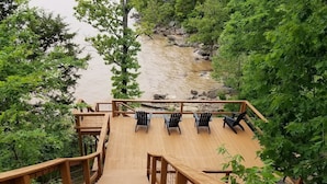 A wooden deck leading to a seating area with chairs, surrounded by greenery and a river. - Lakefront Oasis | Hot Tub | Game Room | 3+ Acres (Stigler)