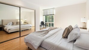 Une chambre moderne avec un grand lit, une porte vitrée donnant sur une autre pièce, un fauteuil et une fenêtre avec des stores.