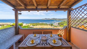 A balcony with a wooden table set for dining, overlooking a coastal view with the sea and distant buildings. - Asfodeli - Flat with sea view and WiFi (Olbia)