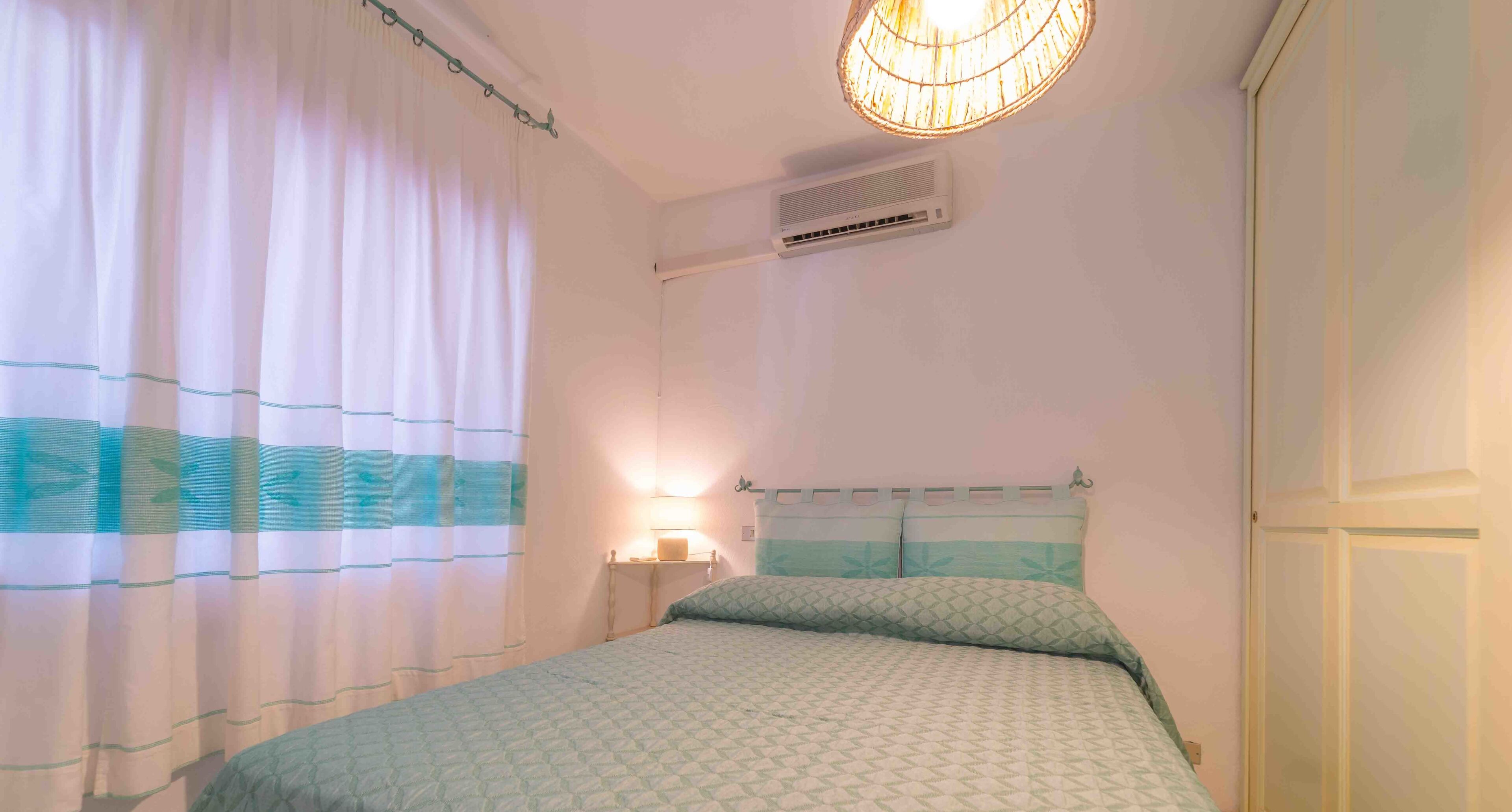Ein Schlafzimmer mit einem Bett, einem Nachttisch mit Lampe, einem Fenster mit Vorhängen und einer Klimaanlage an der Wand.