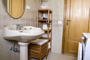 Une salle de bain avec un grand lavabo, une étagère en bois où sont rangés les articles de toilette, et une porte en bois.
