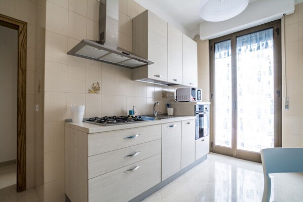 A modern kitchen with a gas stove, oven, and microwave, light-colored cabinets, and a large window with a view. - WePuglia - Albero di Giada - Mare Azzurro (Polignano A Mare)