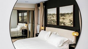 A bedroom with a large mirror reflecting the room, a bed with white linens, a nightstand with a lamp, and a view of a desert landscape through the window.