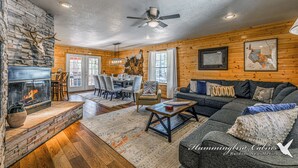 A spacious living room with a stone fireplace, wooden walls, a ceiling fan, and a dining area with chairs and a table. - Mescalero Cabin Pet Friendly | Your Adventure Basecamp w/ Grill, Decks & Cozy Comfort (Alto)