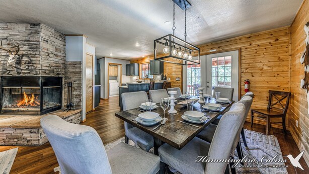 A dining area with a stone fireplace, wooden walls, and a modern chandelier. - Mescalero Cabin Pet Friendly | Your Adventure Basecamp w/ Grill, Decks & Cozy Comfort (Alto)
