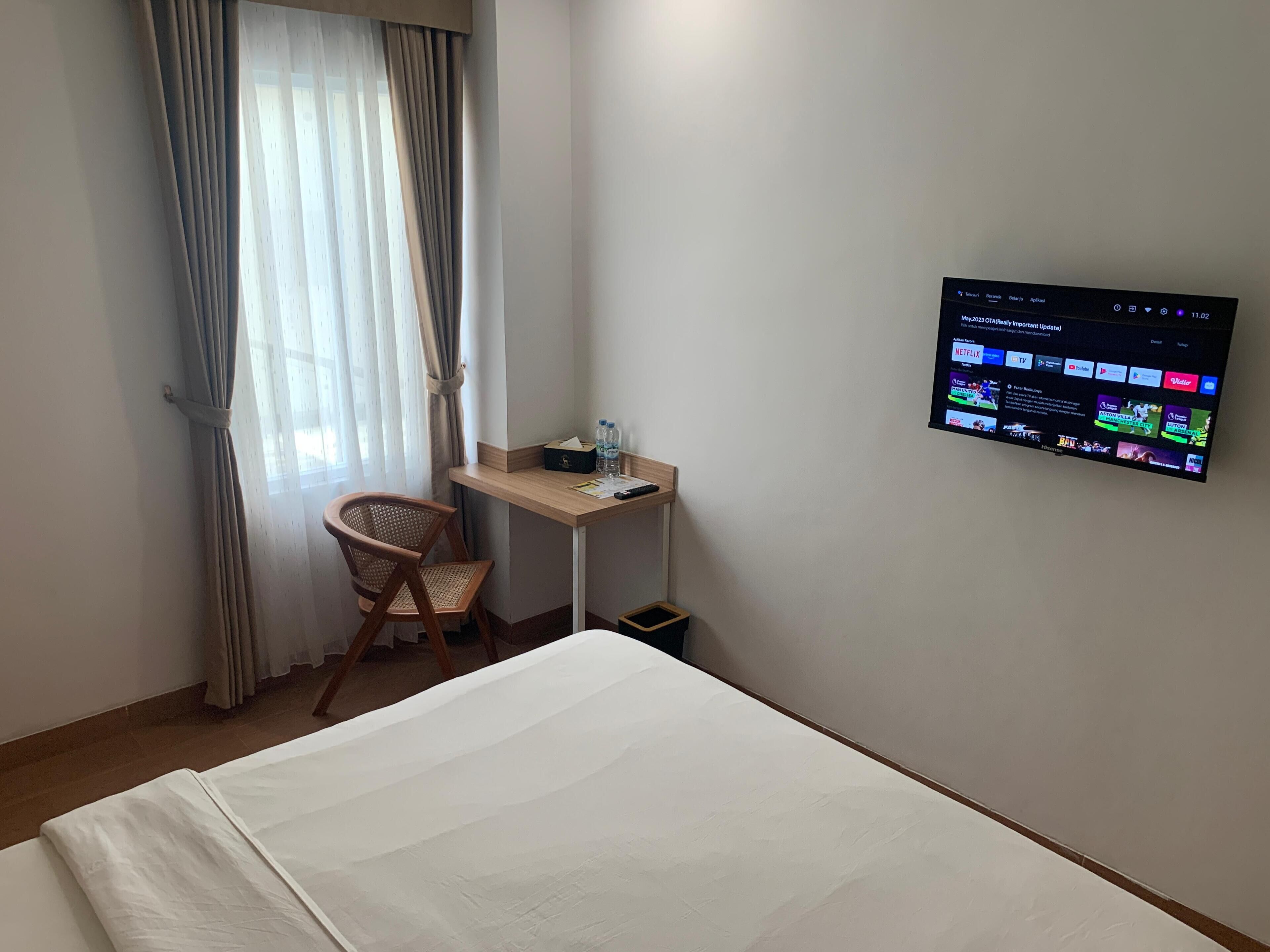 Habitación de hotel con cama, escritorio, silla, televisión montada en la pared y ventana con cortinas.
