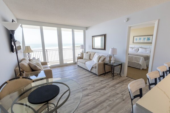 A spacious living room with a glass table, a sofa, and a bed with a balcony view.