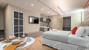 A modern hotel room with a bed, a TV mounted on the wall, a small kitchenette, and a dining area with a table set for two.