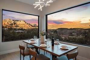 Un coin repas avec une table dressée pour quatre personnes, de grandes fenêtres offrant une vue sur un paysage montagneux au coucher du soleil.