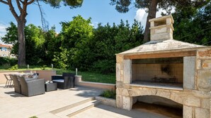 An outdoor patio with a stone-built barbecue area, a dining set, and a view of a lush green landscape.