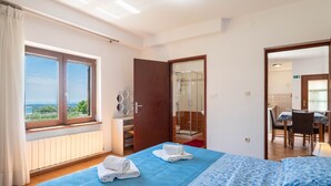 A hotel room with a large bed, a bathroom with a shower, and a dining area visible through an open door.