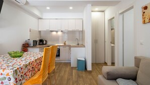 Une cuisine compacte avec des meubles blancs, un four à micro-ondes et un évier. On y trouve une table à manger avec une nappe colorée et deux chaises. Une poubelle est visible près de l’évier.