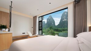 A modern hotel room with a large bed, a desk, a chair, and a view of a mountainous landscape through a large window. - Hidden Dragon Villa (Guilin)