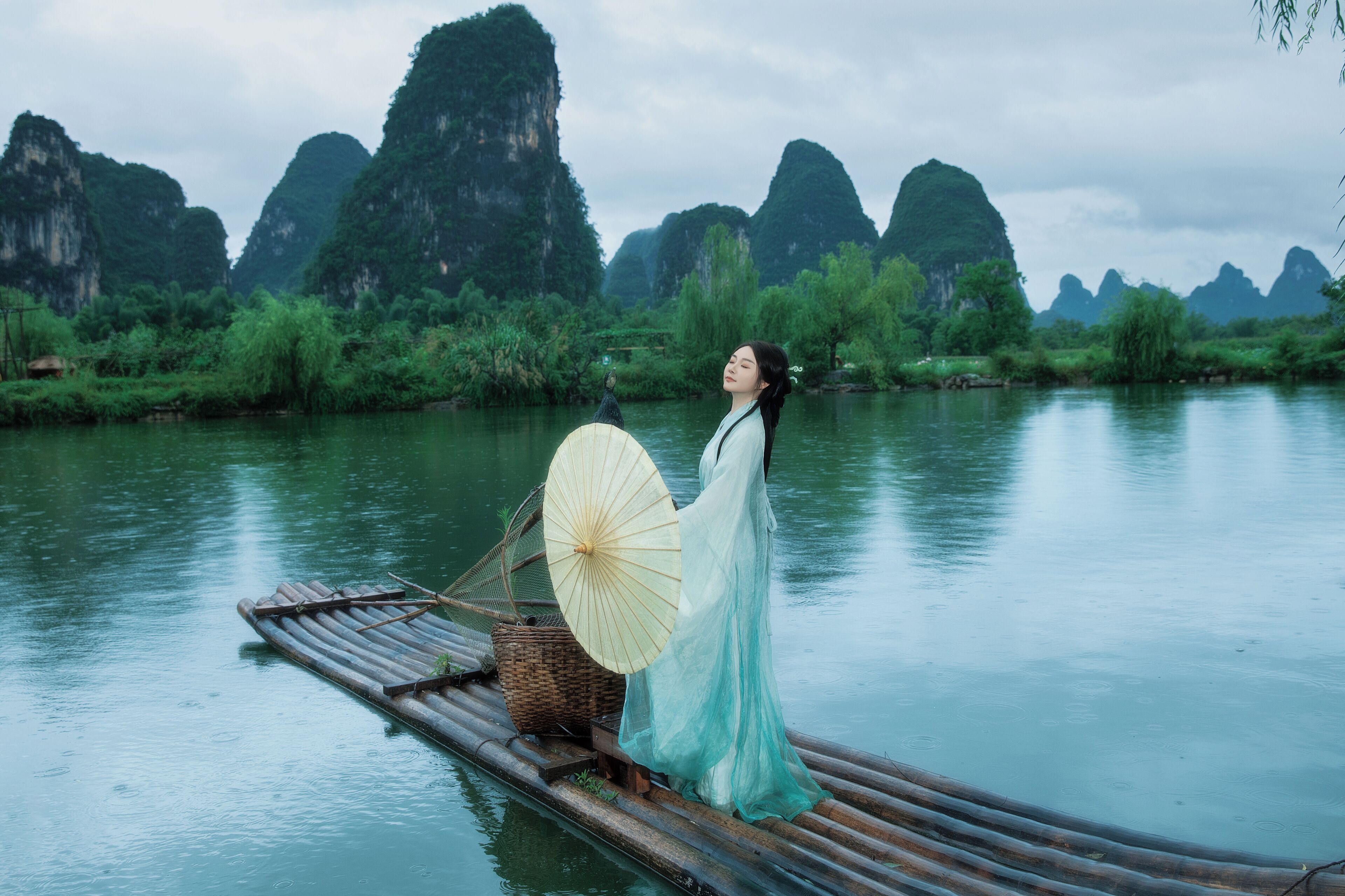 A person on a bamboo raft in a river with green cliffs in the background.