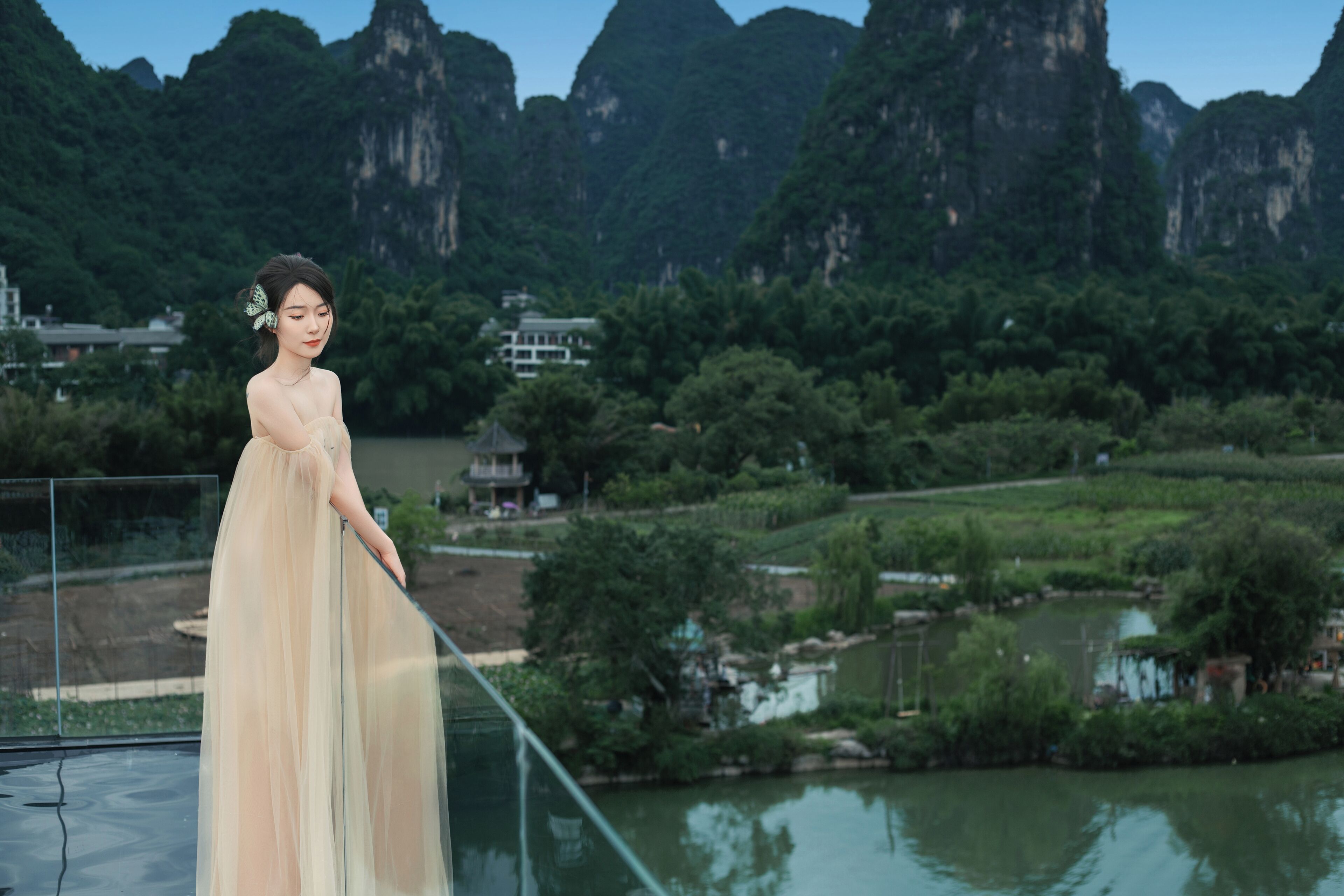 A person in a sheer dress standing by a railing with a view of a river and mountains in the background.