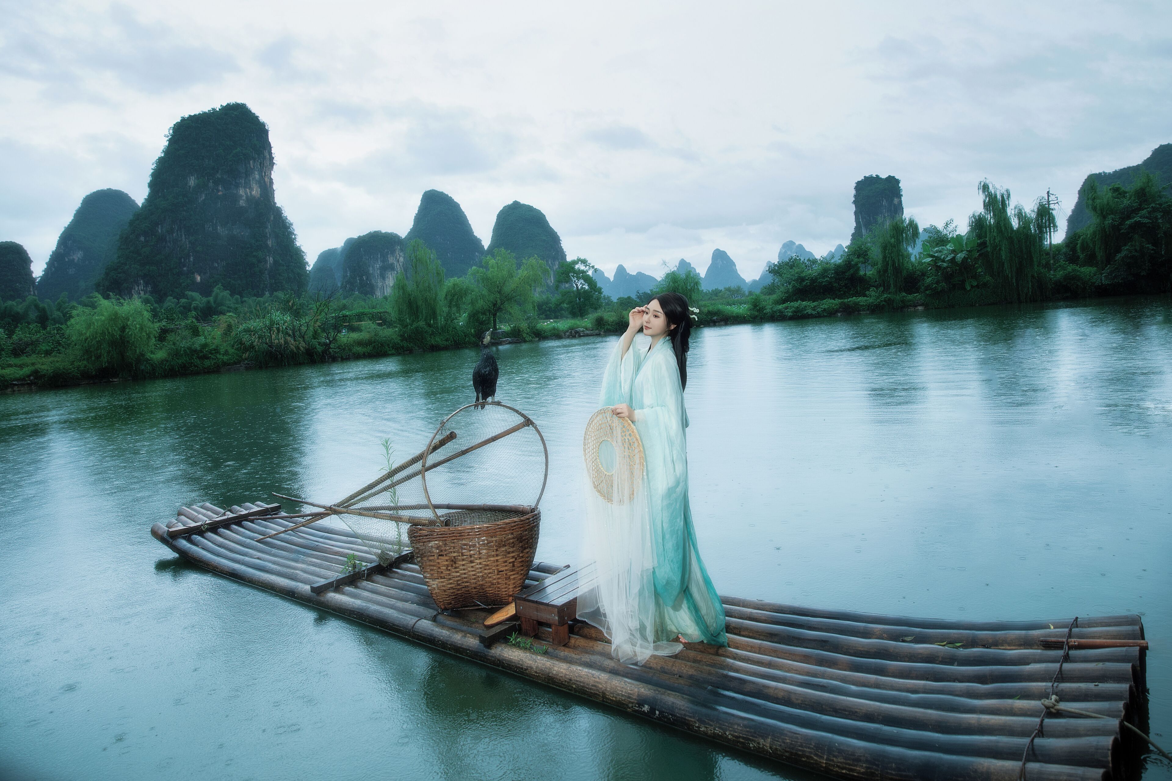 A person in traditional attire standing on a bamboo raft in a river with natural rock formations in the background.
