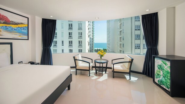 A hotel room with a bed, two chairs, a small table with flowers, a TV, and a large window with a city view.