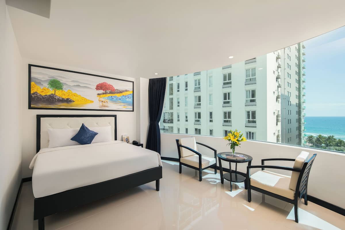 Een hotelkamer met een bed, een stoel, een klein tafeltje met een vaas en een groot raam met uitzicht op gebouwen en de zee.