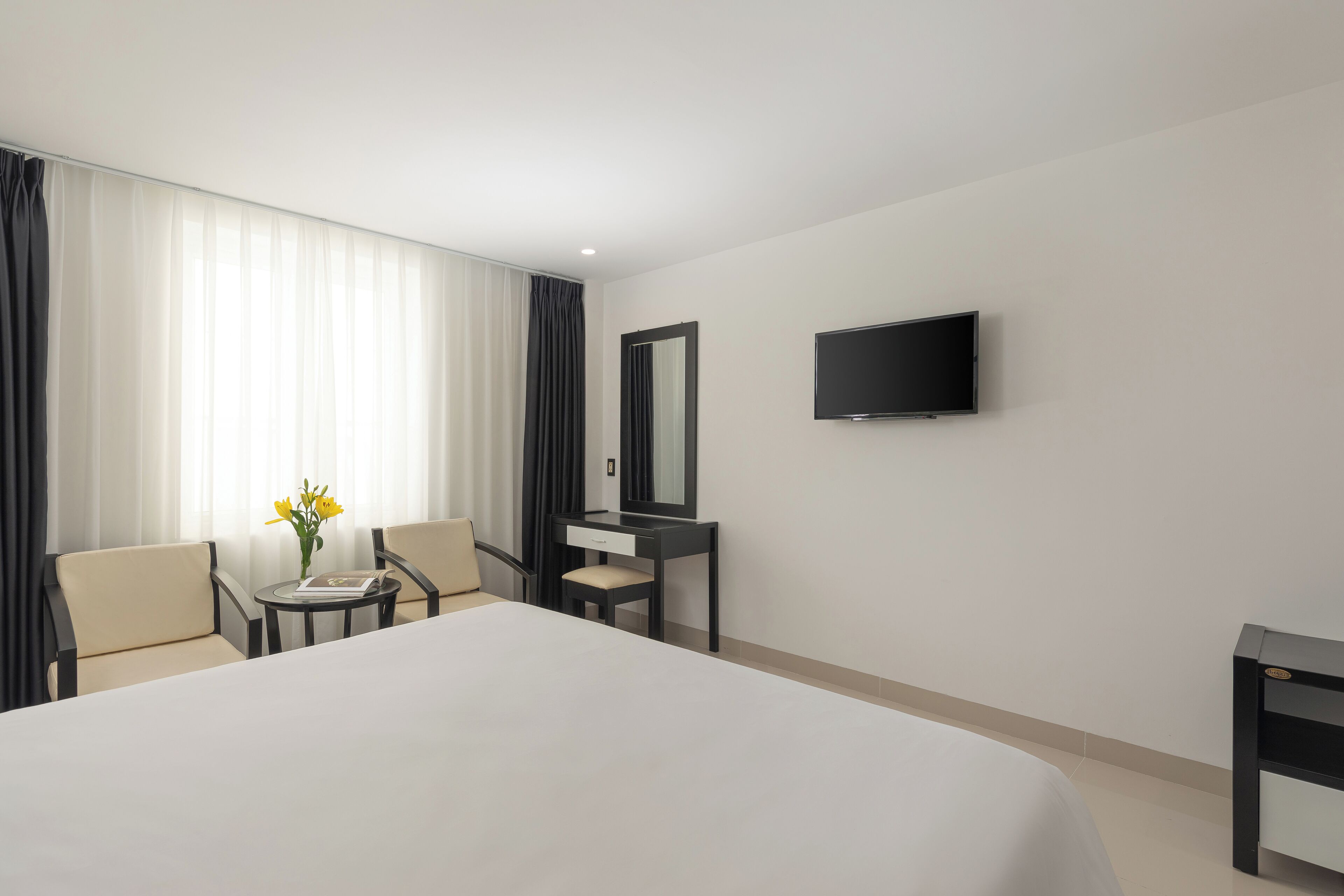 A hotel room with a large bed, a desk with a chair, a small table with a vase of flowers, a mirror, and a TV mounted on the wall.