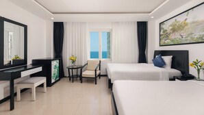 A hotel room with two beds, a desk, a chair, a small table with flowers, a large window with curtains, and a painting on the wall.