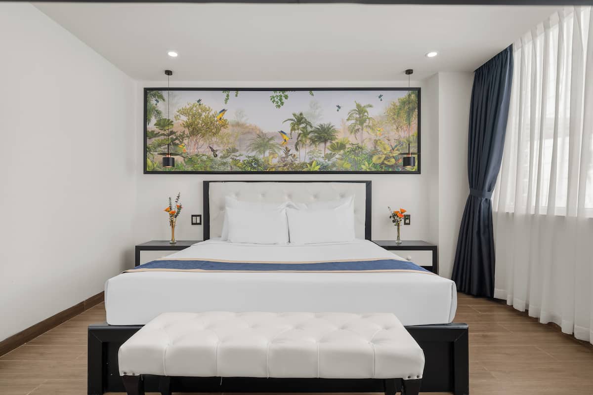 Een moderne hotelkamer met een groot bed, een bankje, een nachtkastje met een vaas, en een schilderij van een tropisch landschap.