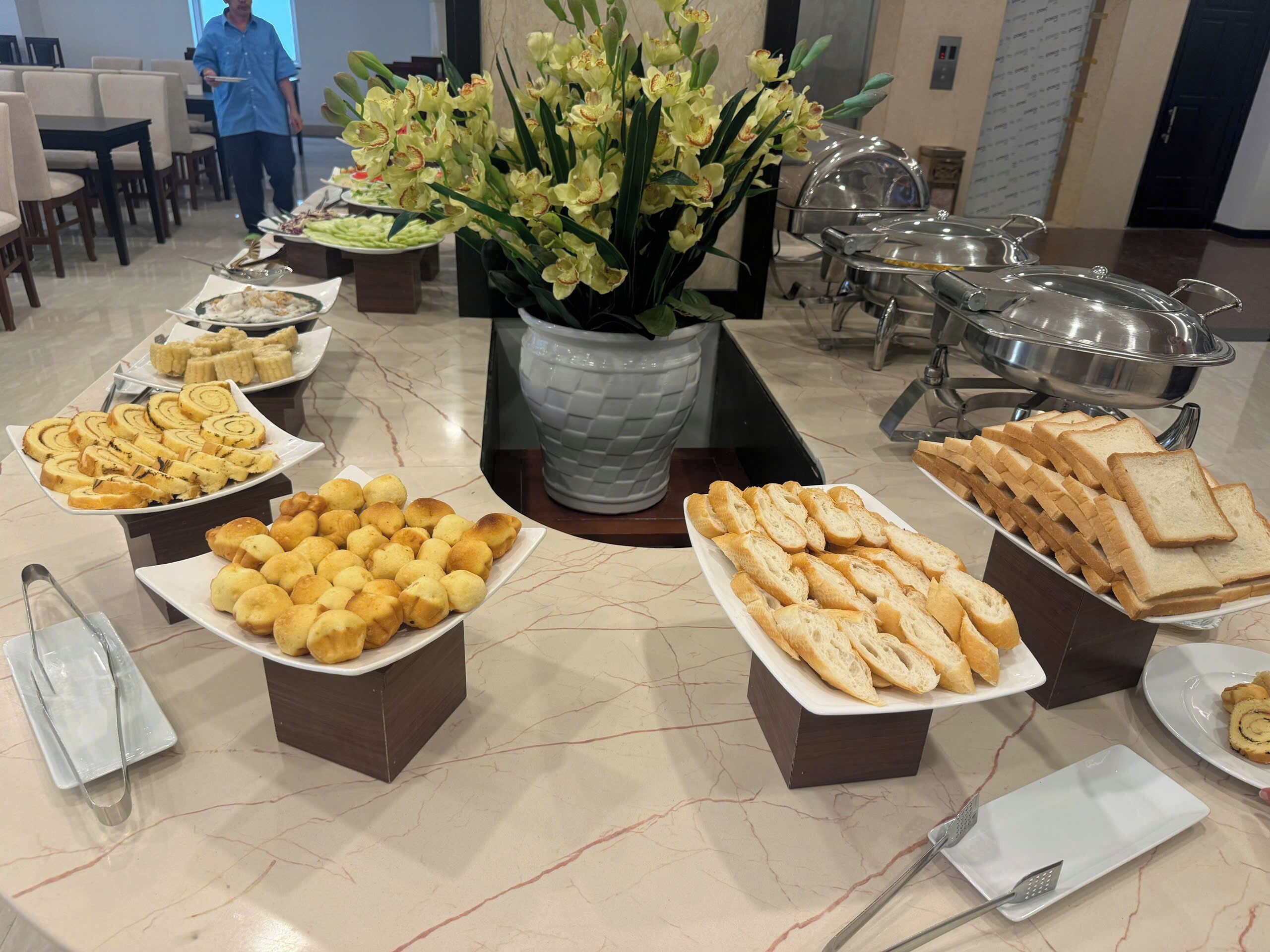 Mesa de buffet con diversos alimentos, una planta en maceta y una persona al fondo.