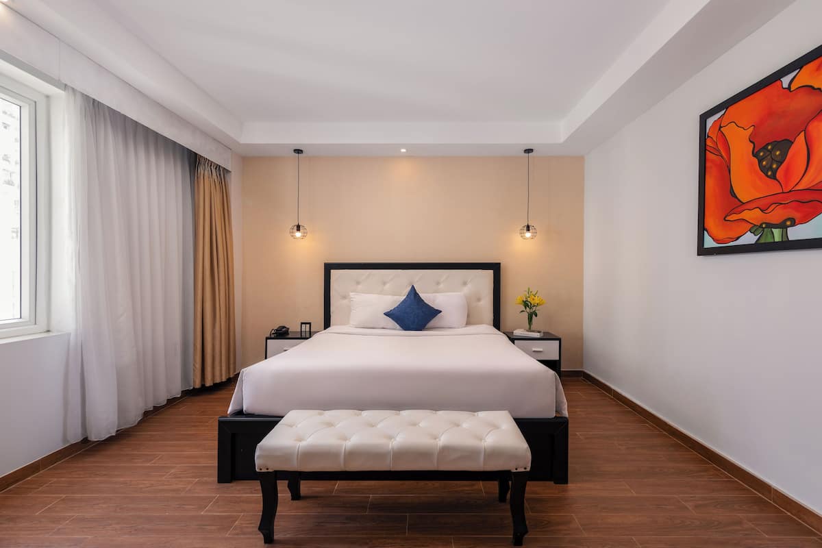 Een slaapkamer met een groot bed, een bankje, een nachtkastje, een wandlamp en een raam met gordijnen.