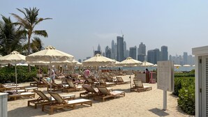 A beach with rows of wooden lounge chairs and beige umbrellas, a city skyline in the background, and a signboard with text. - Unit 146 Seven Palm in Dubai (Dubai)
