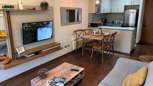 Ein modernes Wohnzimmer mit Flachbildfernseher, Couchtisch aus Holz und einer Couch mit Kissen. Im Hintergrund ist eine Küchenzeile mit Kühlschrank und Esstisch zu sehen.